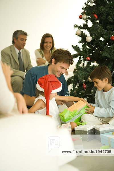 Mann und zwei Kinder öffnen Weihnachtsgeschenke vor dem Weihnachtsbaum  Familie beobachtet im Hintergrund