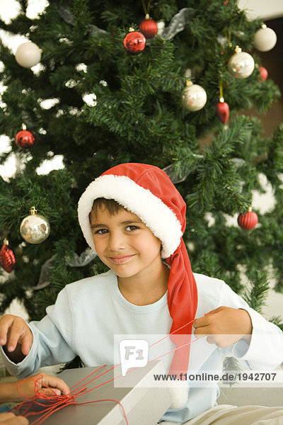 Junge eröffnet Weihnachtsgeschenk vor dem Weihnachtsbaum  mit Weihnachtsmütze  lächelnd vor der Kamera