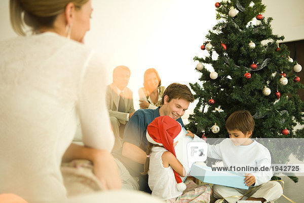 Mann und zwei Kinder eröffnen Weihnachtsgeschenk vor dem Weihnachtsbaum  Frau schaut im Vordergrund zu