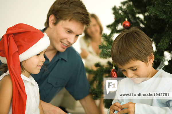 Vater und zwei Kinder eröffnen Weihnachtsgeschenk vor dem Weihnachtsbaum  Mädchen mit Weihnachtsmütze
