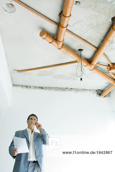 Mann im Anzug  der unter nackten Rohren im Hausinneren steht  mit Telefon und Haltepapier