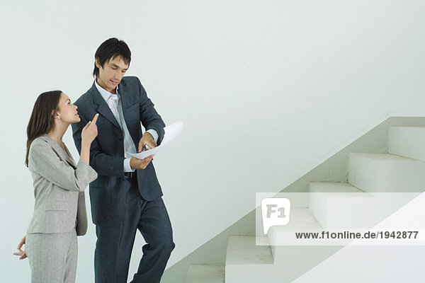 Mann und Frau in Anzügen stehen am Fuß der Treppe  Mann schaut auf Dokument  während Frau nach oben zeigt.