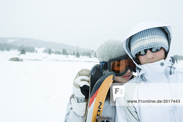 Two young skiers standing side by side  looking at camera  one's head on the other's shoulder