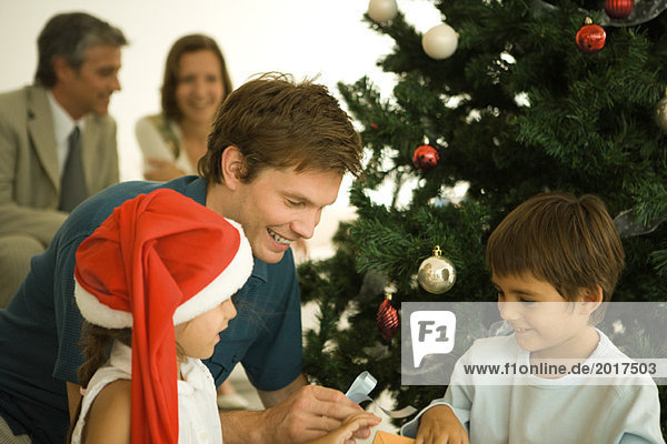 Vater und zwei Kinder am Weihnachtsbaum sitzend  gemeinsam Geschenke öffnen  Tochter mit Weihnachtsmütze