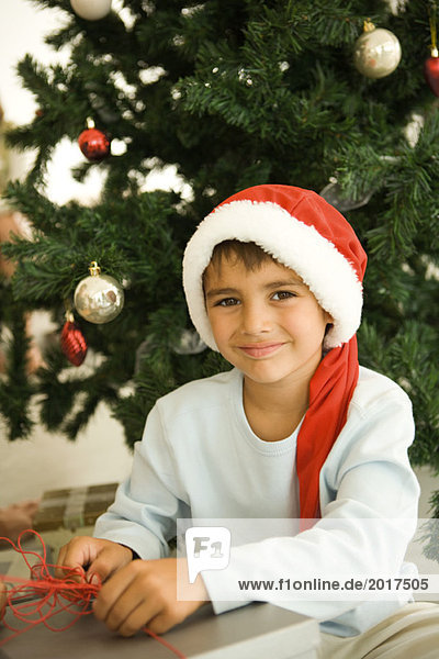 Eröffnungsgeschenk vor dem Weihnachtsbaum  mit Nikolausmütze  lächelnd vor der Kamera