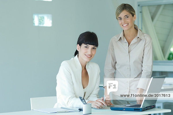 Two young businesswomen in office  one seated at desk  the other standing by her side  both smiling at camera  portrait