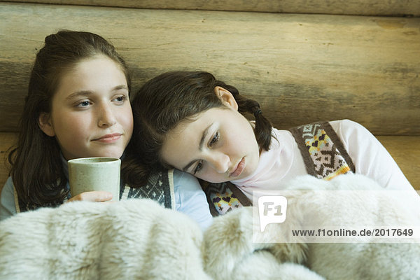 Two teen girls in winter clothes sitting under warm blanket together  one holding hot beverage