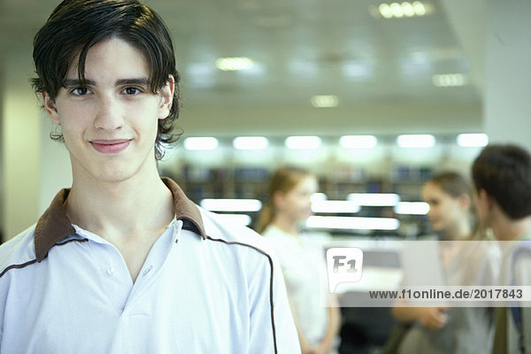 Male college student in library  smiling at camera  other students chatting in blurred background