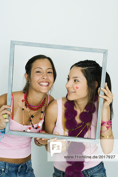 Two young friends holding up frame together  one smiling at camera  portrait