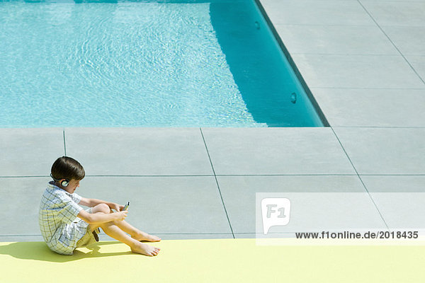Boy sitting on the ground next to swimming pool  using handheld electronic device  wearing headphones