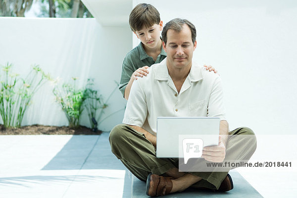 Man sitting on ground using laptop computer  son looking over his shoulder