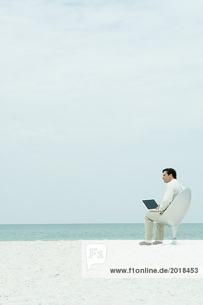 Geschäftsmann sitzt auf einem Stuhl am Strand  benutzt einen Laptop und schaut weg.