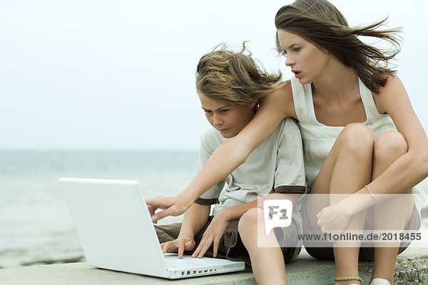 Teen girl and little brother using laptop computer together  girl pointing over boy's shoulder