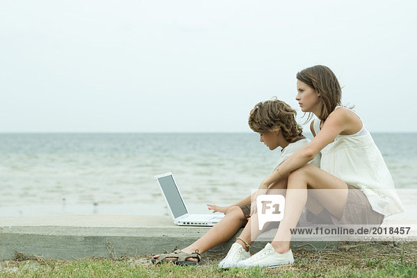 Teenager-Mädchen und kleiner Bruder mit Laptop-Computer zusammen am Meer  Seitenansicht