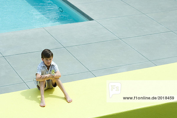 Boy sitting on the ground by swimming pool  looking at cell phone