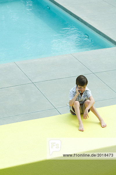 Boy sitting on the ground in front of swimming pool  using cell phone  looking down