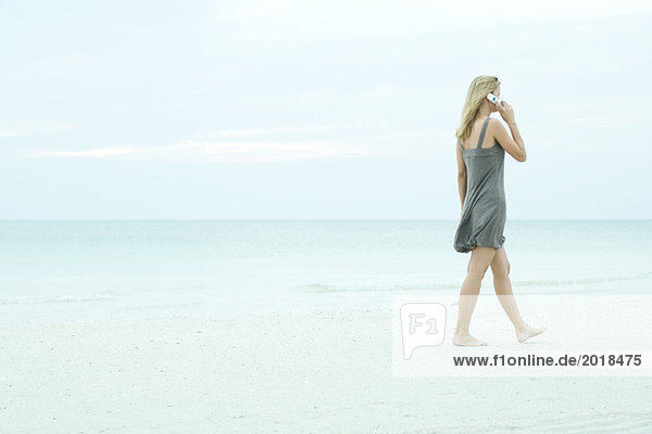 Junge Frau  die am Strand spazieren geht  mit dem Handy  volle Länge