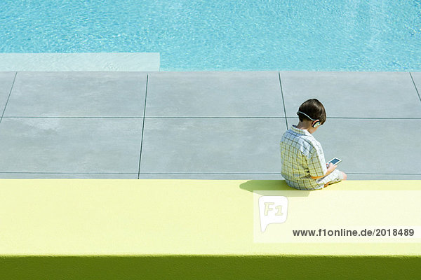Boy sitting by swimming pool  using handheld electronic device  wearing headphones  rear view