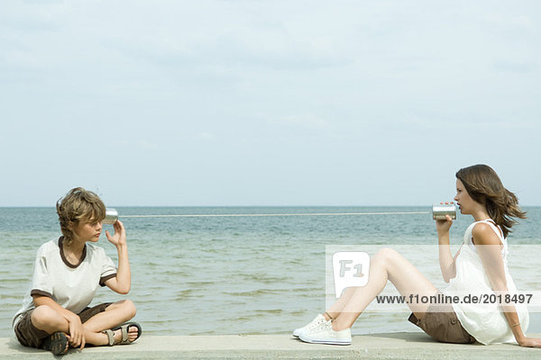Junge und Teenager-Mädchen sitzen in der Nähe des Ozeans und reden durch die Blechdose.