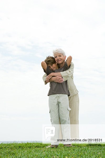 Seniorin und Enkelin auf Gras stehend  umarmend  lächelnd vor der Kamera  volle Länge
