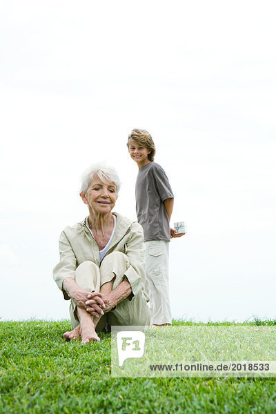 Seniorin auf Gras sitzend  Enkelin hinter ihr stehend  Geschenk haltend  lächelnd  volle Länge