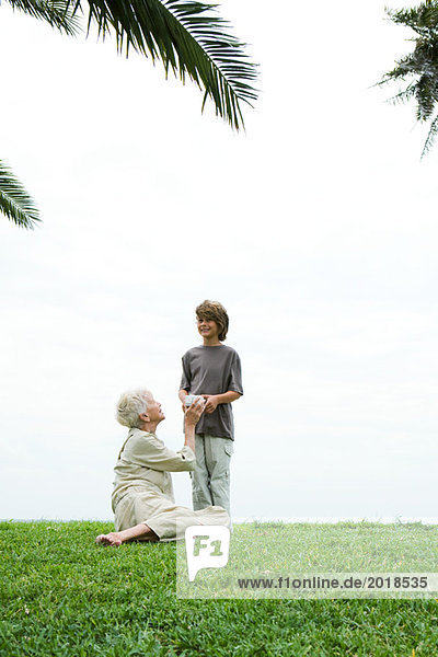 Seniorin auf Gras sitzend  Enkelin bei der Übergabe ihres Geschenks  volle Länge