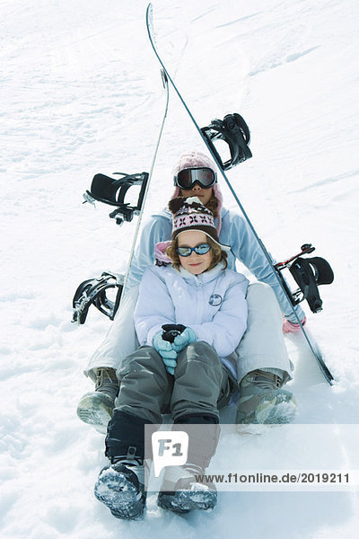 Two young skiers sitting under skis together  smiling at camera  portrait