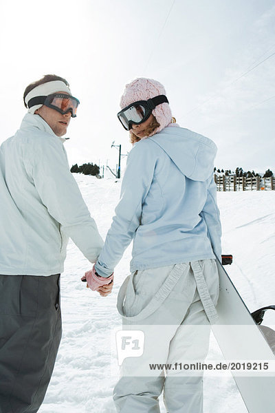 Zwei junge Freunde gehen,  Händchen halten,  über die Schultern schauen,  einer trägt Snowboard,  Rückansicht