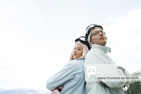 Teen girl and young man in ski gear  standing back to back  arms folded  smiling  girl looking at camera