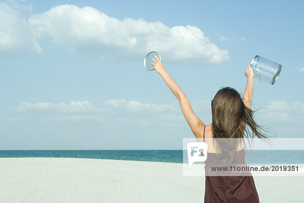 Frau steht am Strand  Rückansicht  hält klaren Behälter hoch  fängt Wolken auf  optische Täuschung