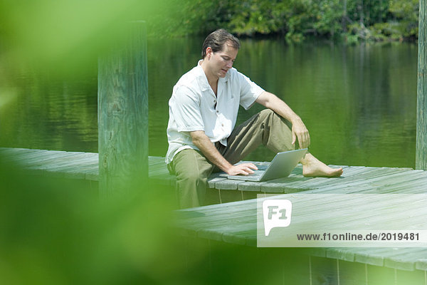 Man sitting on dock using laptop computer  view through foliage