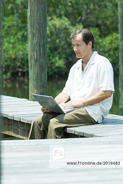 Mann sitzt auf dem Dock  benutzt den Laptop und schaut weg.