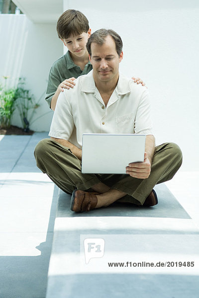 Man sitting on ground using laptop computer  son looking over his shoulder