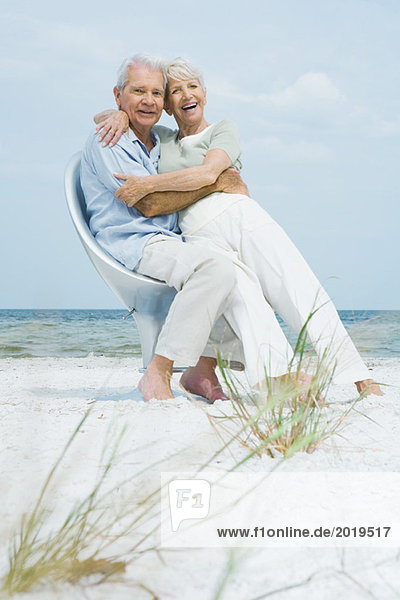 Seniorenpaar sitzt zusammen auf einem Stuhl am Strand  umarmt  Frau sitzt auf dem Schoß des Mannes  beide lächelnd vor der Kamera.
