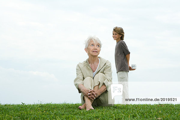 Seniorin auf Gras sitzend  Enkelin hinter ihr stehend  Geschenk haltend  Kamera lächelnd  volle Länge