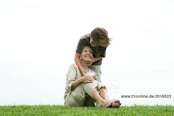 Seniorin auf Gras sitzend  Enkelin steht hinter ihr mit der Hand auf dem Gesicht und gibt ihr ein Geschenk.