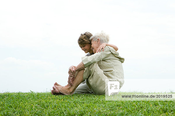 Seniorin und Enkelin  sitzend auf Rasen  volle Länge