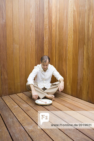 Man sitting with legs crossed on wooden floor  looking down at miniature rock garden  full length