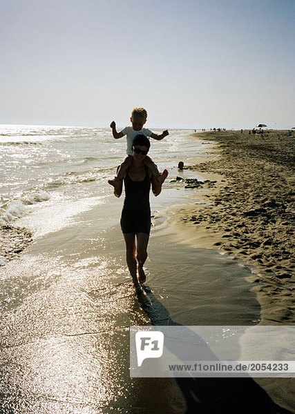 A woman carrying a young boy on her shoulders whilst walking along a beach