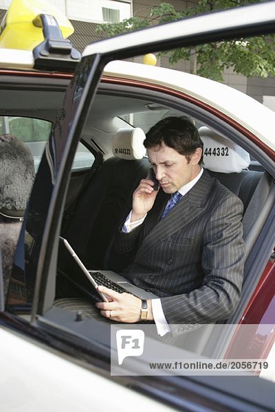 Ein englischer Geschäftsmann telefoniert und arbeitet am Laptop während er auf dem Rücksitz eines deutschen Taxis sitzt