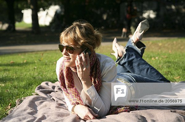 Eine junge Frau liegt auf einer Decke im Park und telefoniert