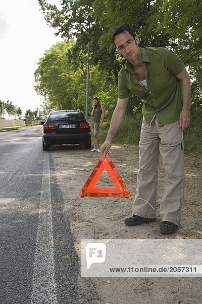 Ein Mann stellt ein Warnungsdreieck am Straßenrand auf  sein Auto und eine Beifahrerin im Hintergrund