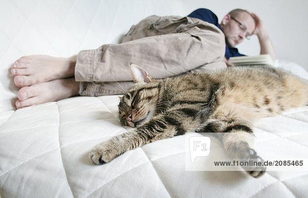 Close-up of cat sleeping with man reading book on bed