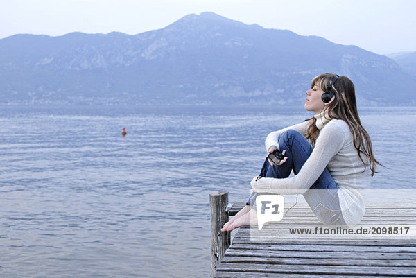 Italy  Lake Garda  Young woman (20-25) sitting on jetty holding personal stereo