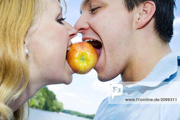 Porträt eines jungen Paares  das auf einen Apfel zwischen den Mündern beißt.