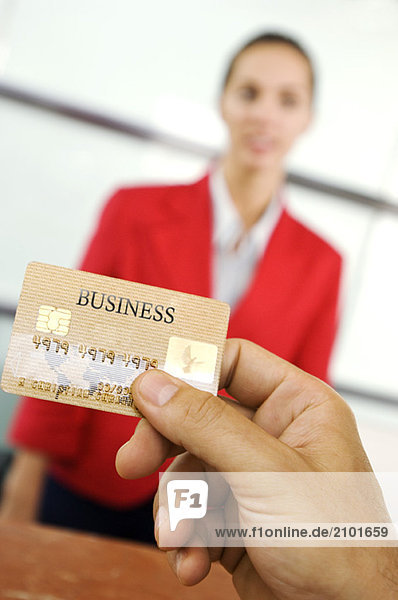 Human hand holding credit card with businesswoman standing in background  close-up