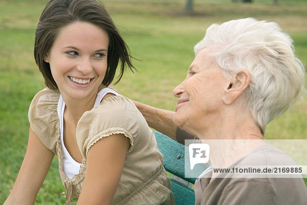 Großmutter und jugendliche Enkelin sitzen zusammen im Freien und lächeln sich an.