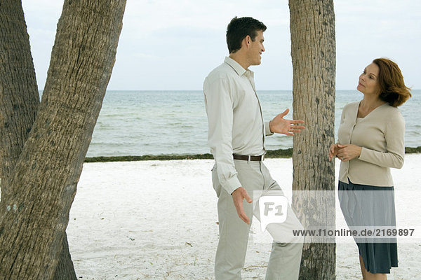 Mann und Frau stehen neben einem Baumstamm am Strand und lächeln sich an.