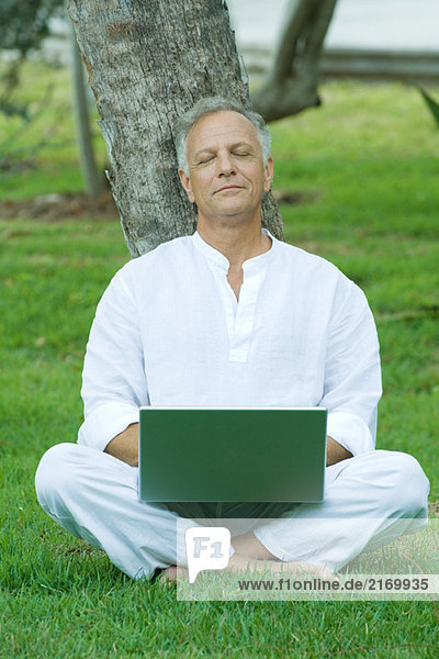 Mature man sitting in grass with laptop computer on lap  eyes closed