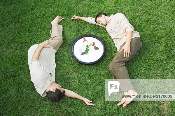 Two men lying on grass around bowl with flower  eyes closed
